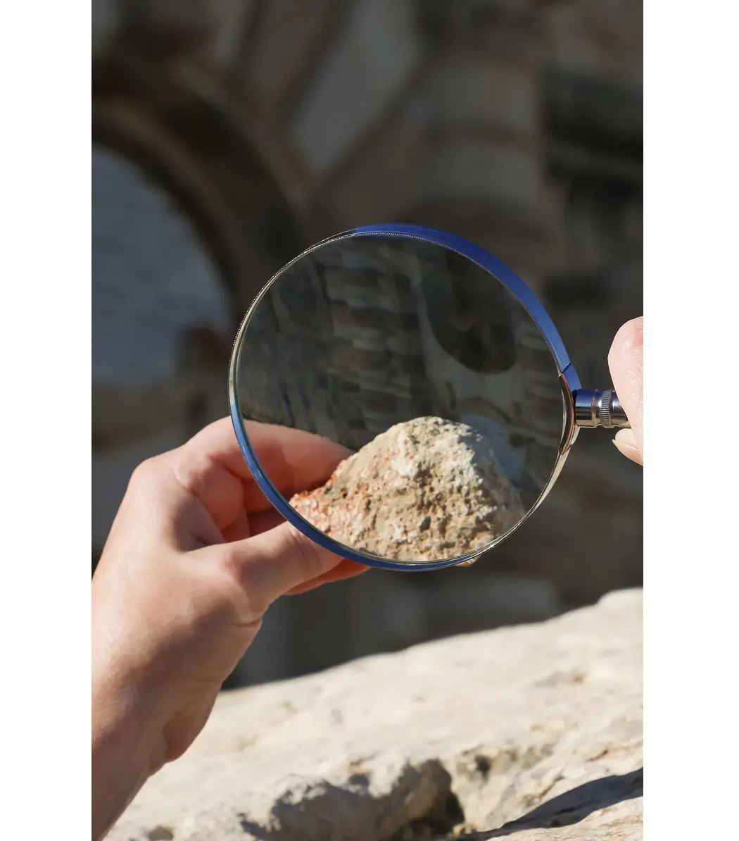 Person examining sand through a magnifying glass outdoors.